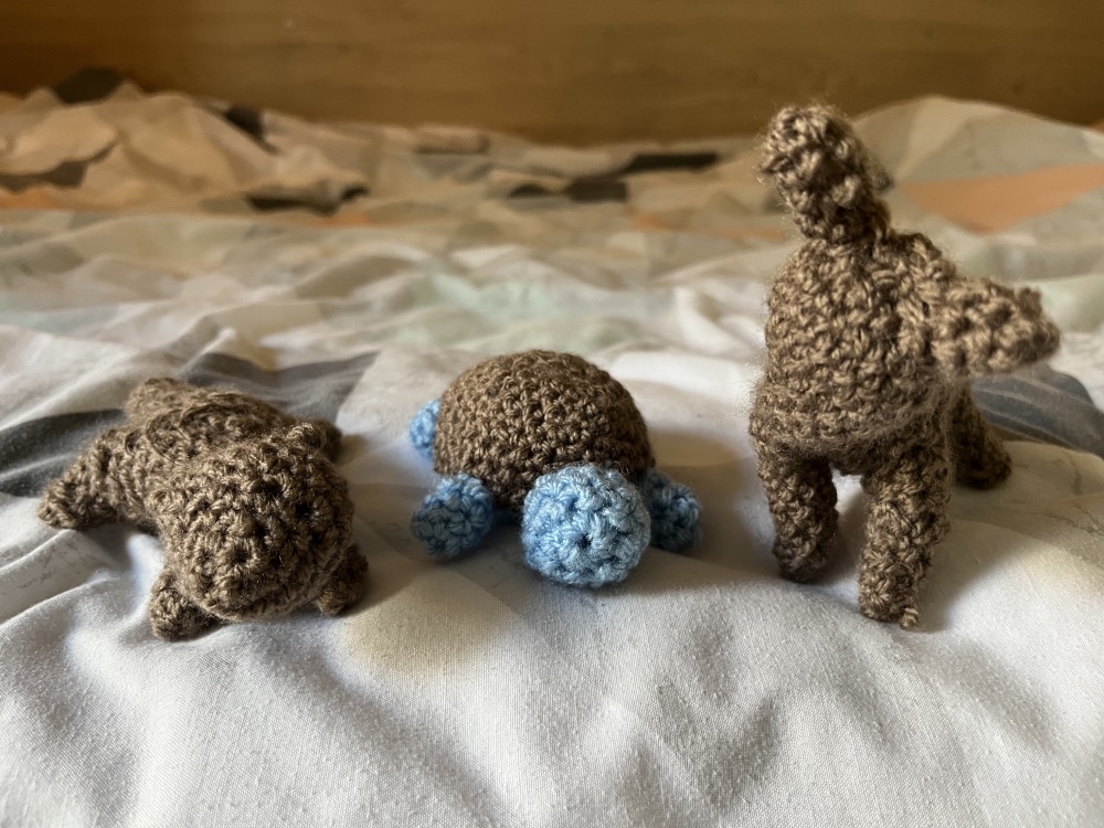 The first three amigurumi animals I’ve made, all placed on the duvet. From left to right: a brown otter with its short limbs and big tail sprawled out like a chubby starfish; a round blue turtle with a brown shell facing forward; and a shy brown deer with unsteady limbs posing with its head drooping towards its chest.