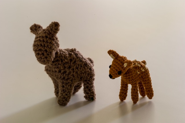 Photo of two crocheted deer placed side by side against a white background. The one on the left is a dark brown deer with noticeably larger gaps between the stitches. The one on the right is a smaller deer with a similar standing posture, except it’s in light brown with white spots across its body. It has safety eyes inserted and an embroidered nose and mouth.