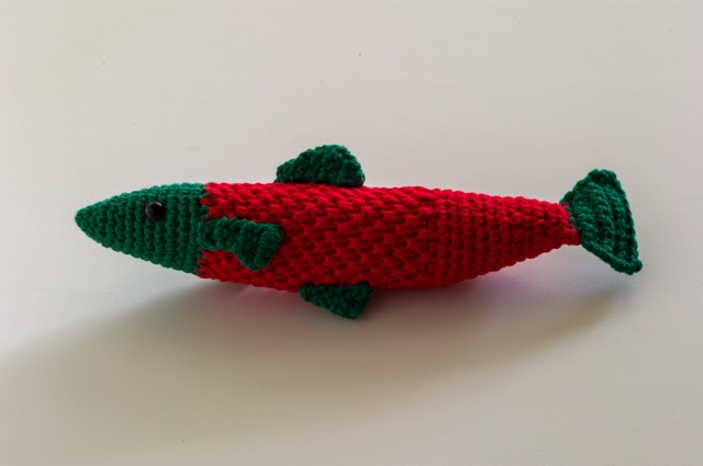 Photo of a crocheted salmon plush against a white background. The salmon has a head and fins in dark green and a red body so it resembles a sockeye salmon.