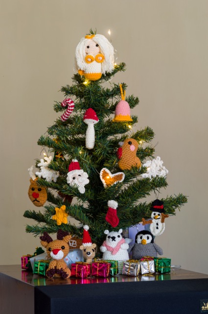 Photo of a tree, about 30cm tall, on a wooden surface. On the tree hangs various crocheted decorations all made from the advent calendar, surrounded by mini fairy lights. Visible from the top to bottom, an angel with messy white hair, a candy cane, a pink bell with yellow lining, a mushroom, two snowflakes, a robin, Santa's face, a gingerbread heart, a reindeer, a star, a stocking, and a snowman. Beneath the tree sits toys from the calendar that are too big for the tree. From left to right, Rudolph, a teddy bear pencil cap, a polar bear, and a penguin. Many tiny presents in foil wrapping surround them.