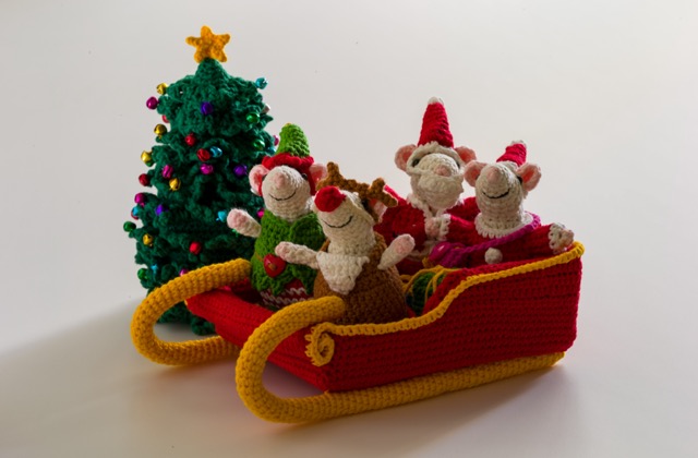 Photo of four crocheted mice sitting in a red sleigh, behind them is a Christmas tree with colourful bells hanging.