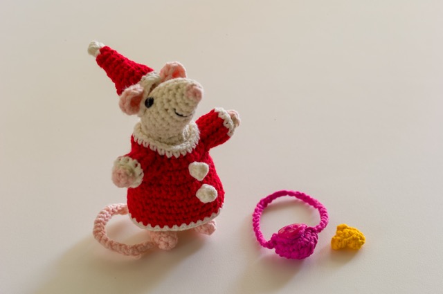 A smiling cream coloured mouse, wearing Mrs Claus' dress and a matching hat. Next to her lies a pink handbag with a button attached, and a piece of cheese.