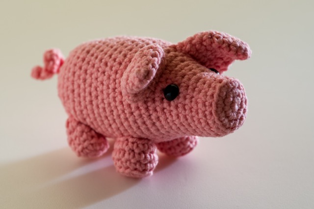 Photo of a pink crocheted pig with a curious expression in a plain background.